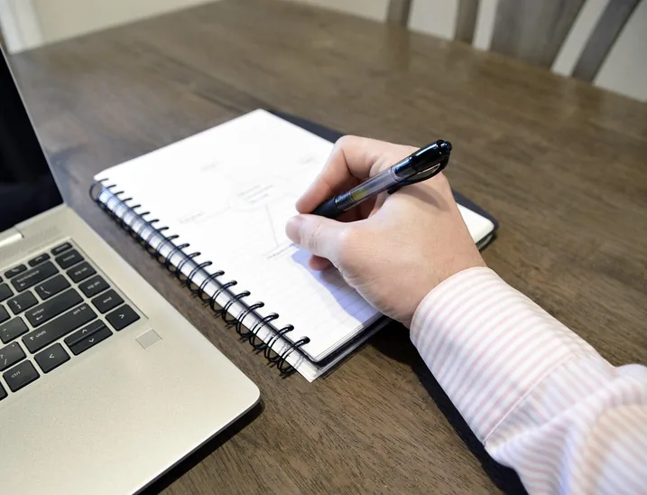 A hand writes the next line in a notebook beside an open laptop, the way an owner works out what a purchase cost.