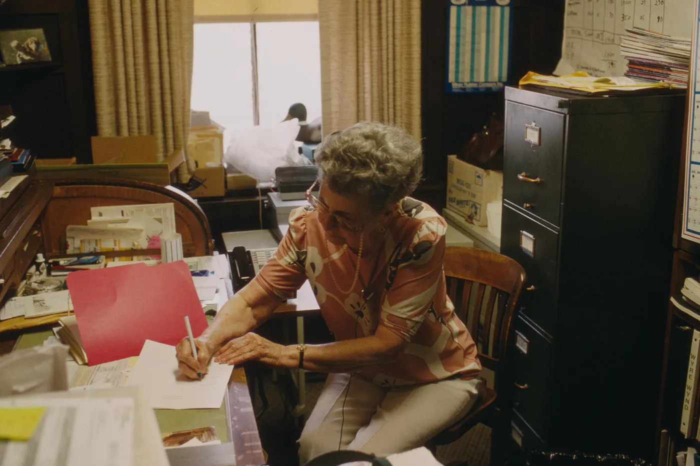 A woman writing on a stack of papers beside a file cabinet and a wall calendar.