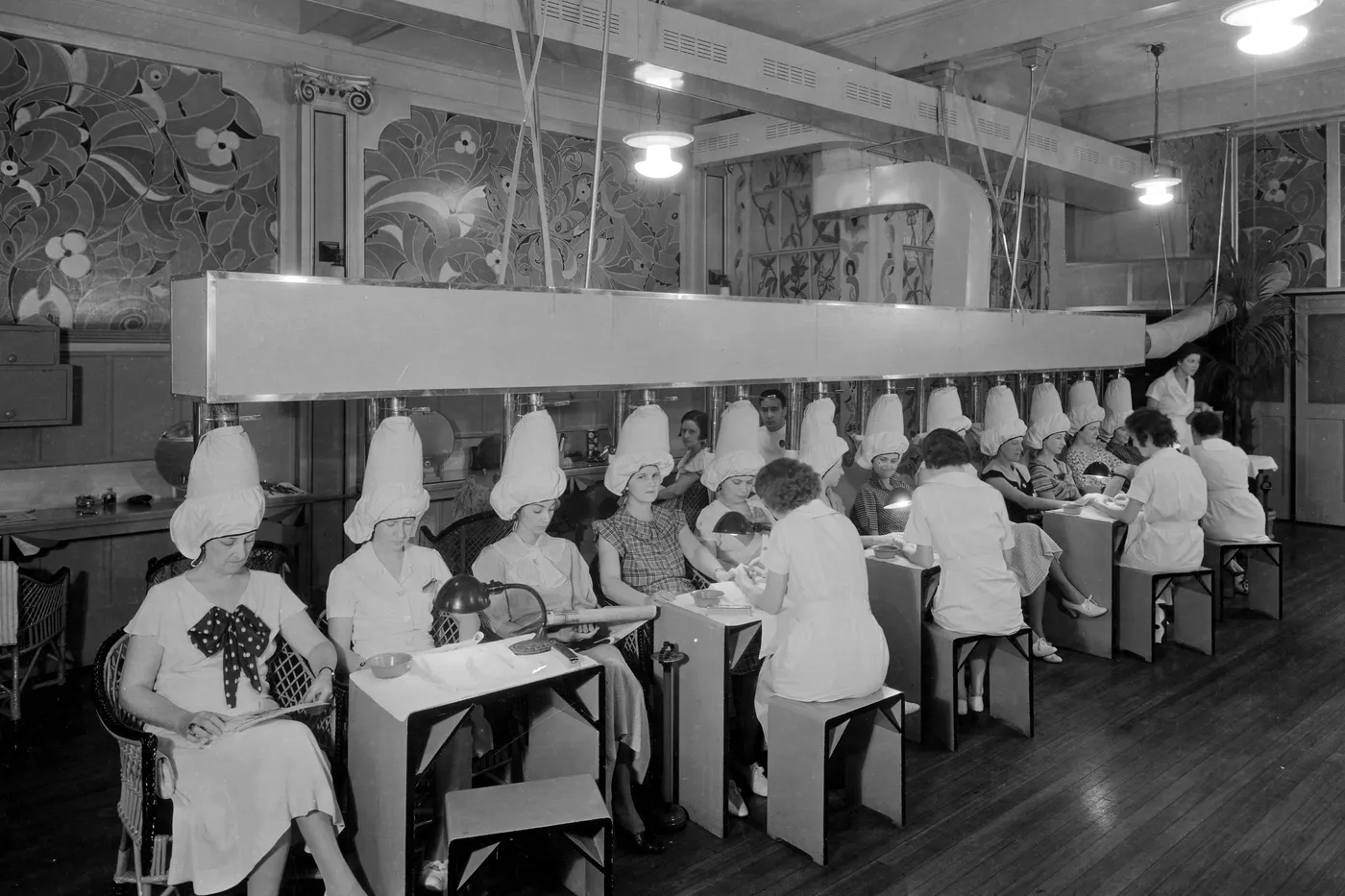 A shop floor of dryer chairs and manicure tables, with workers and guests.