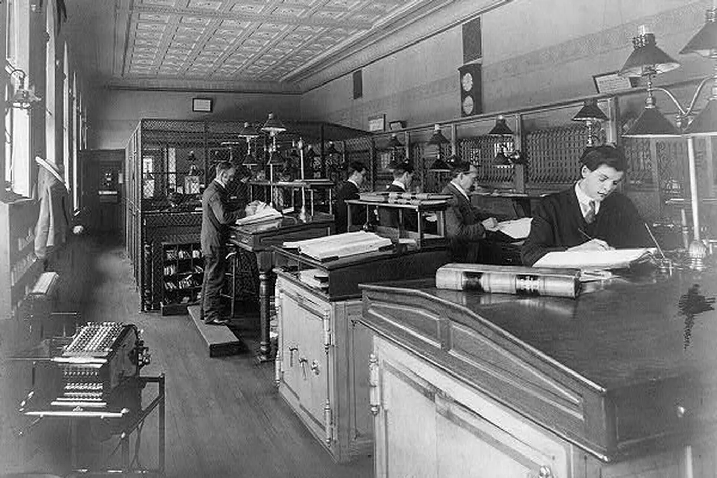 Clerks writing in ledgers along a row of cages, with an adding machine in front.