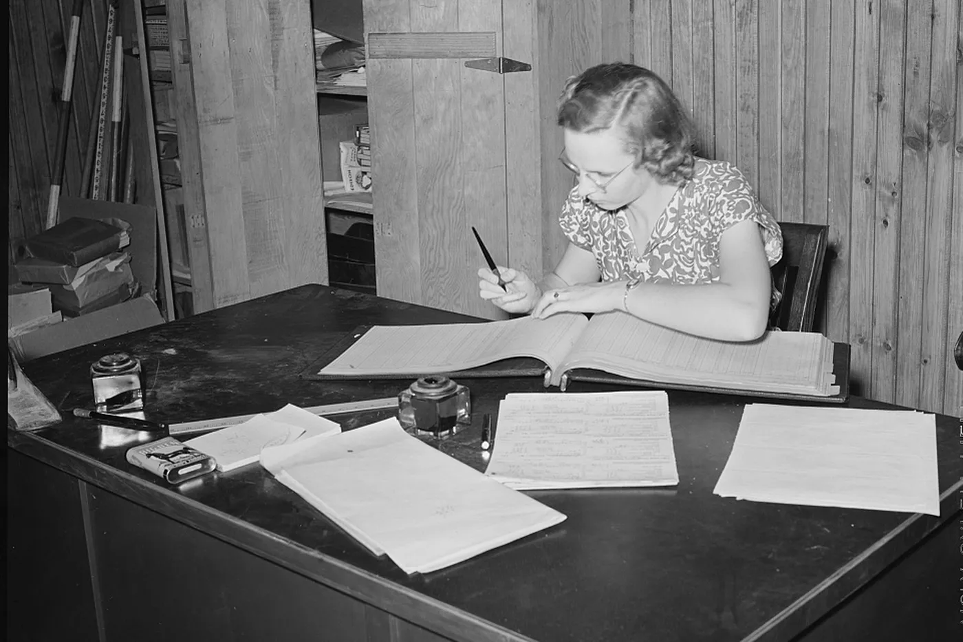 A bookkeeper writing in an open ledger, with loose sheets spread on the table.