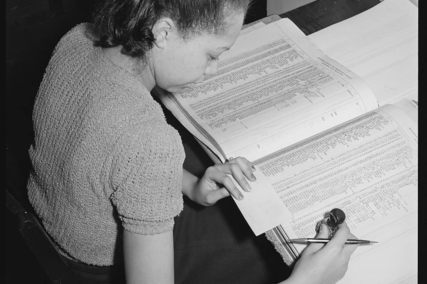 A bookkeeper reading a long ledger, with an adding machine beside the page.