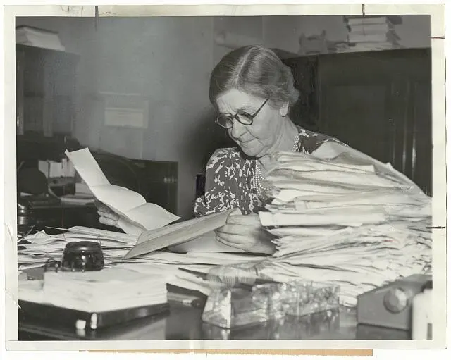 A woman works through a table piled with papers.