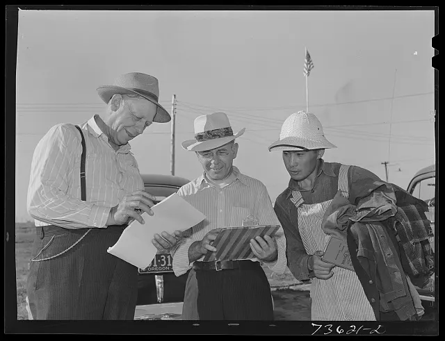Three men read one sheet of paper together outdoors.