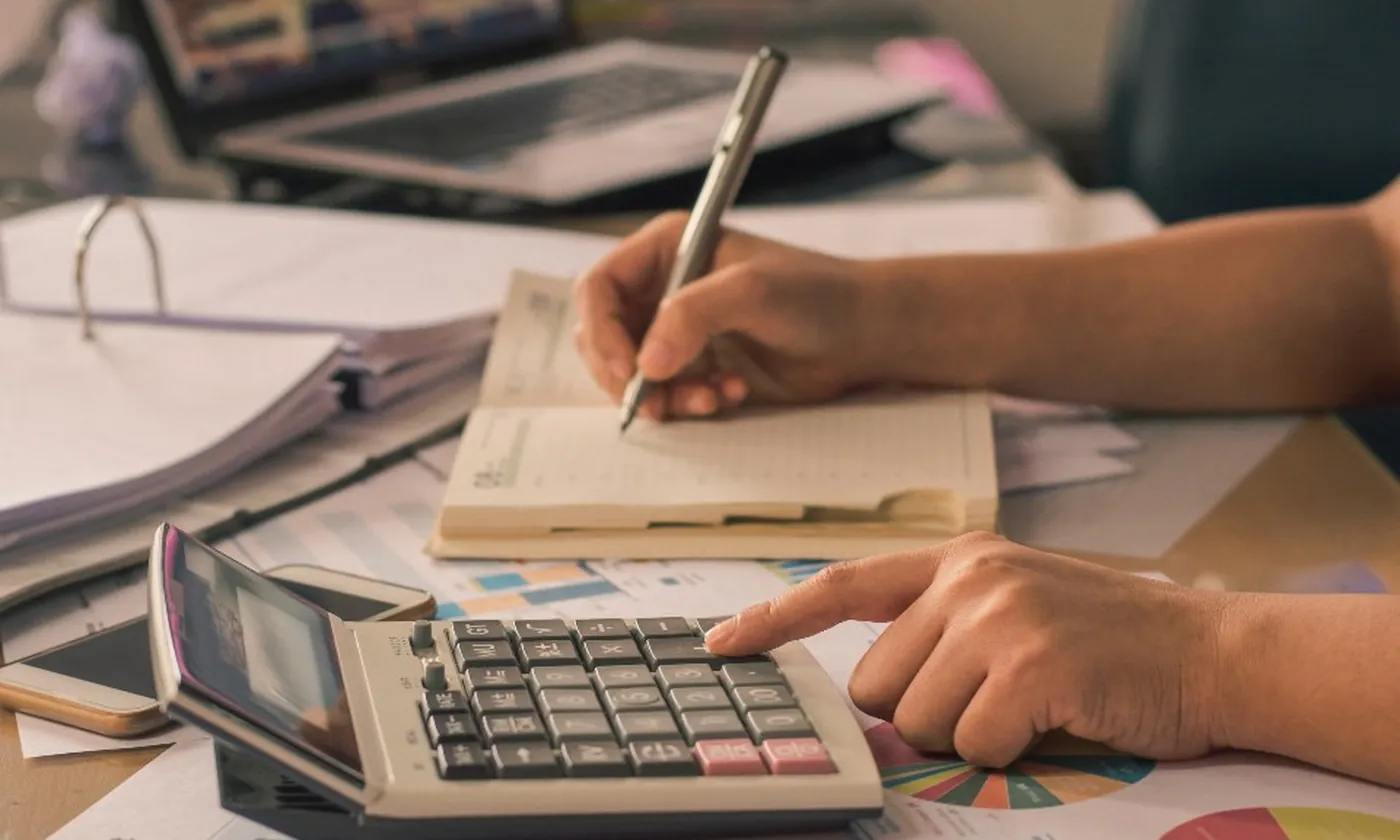 A hand writing figures on a sheet beside a calculator and a binder