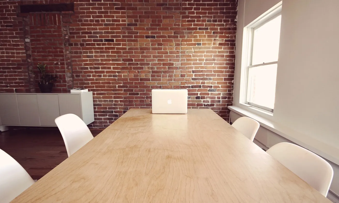 A long table with empty chairs and one laptop beside a window