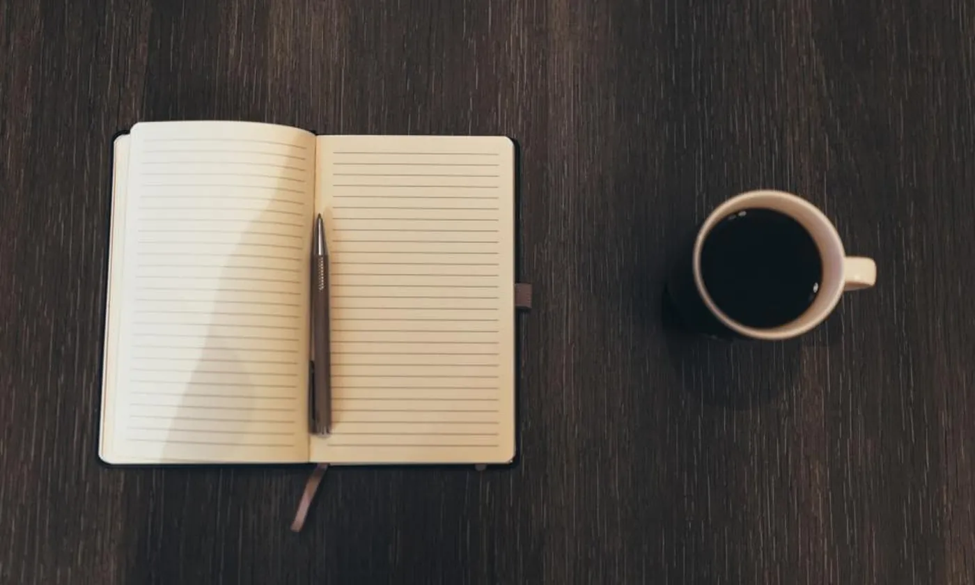 An open notepad with a pen beside a cup of black coffee