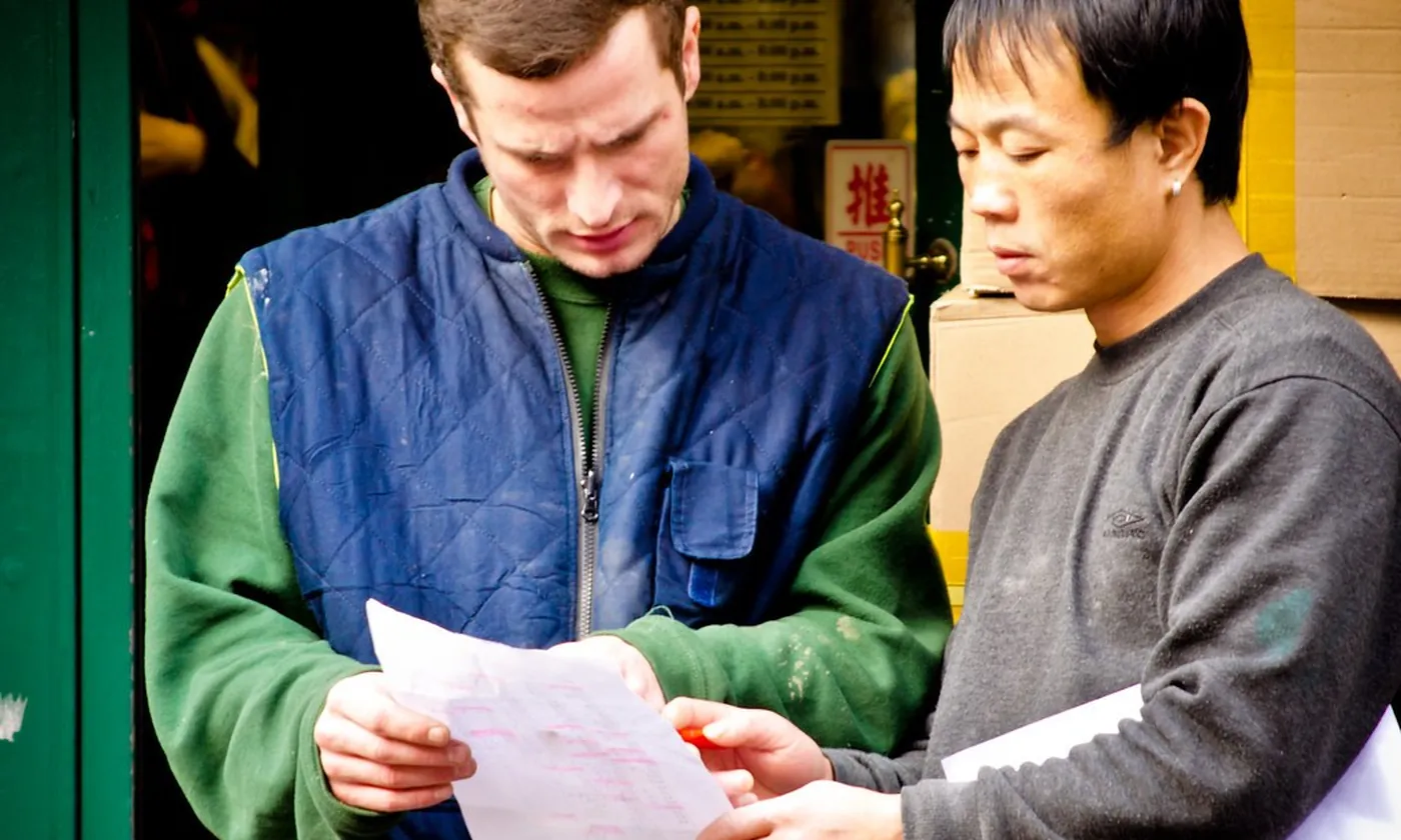 Two men reading a printed order beside a stack of boxes