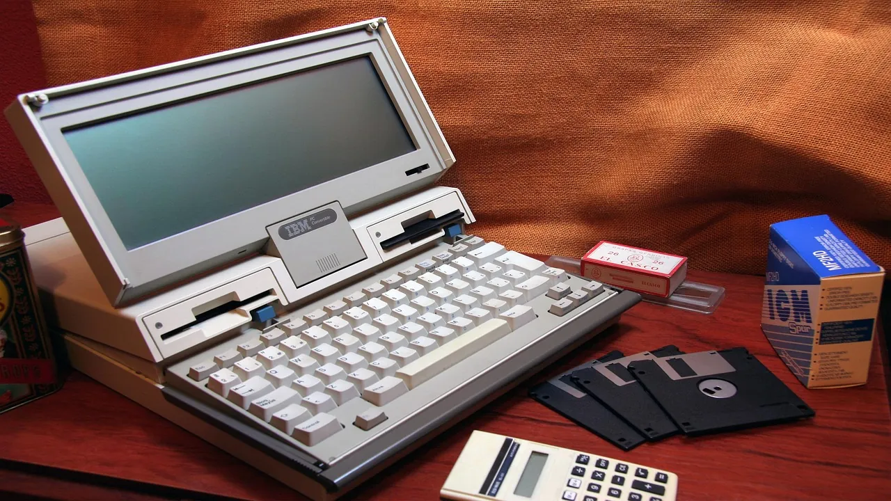 An older laptop beside a calculator and a stack of floppy disks