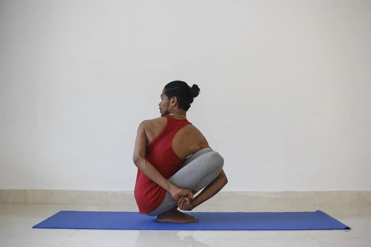 A person holding a seated yoga pose on a blue mat in a plain studio.