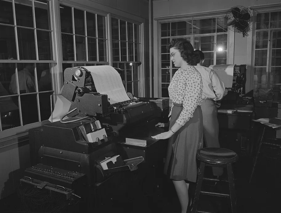 A woman runs a payroll machine. A long sheet of figures feeds through it.