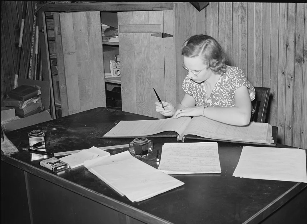A bookkeeper writes in a wide ledger at a wooden desk, with loose sheets beside the book.