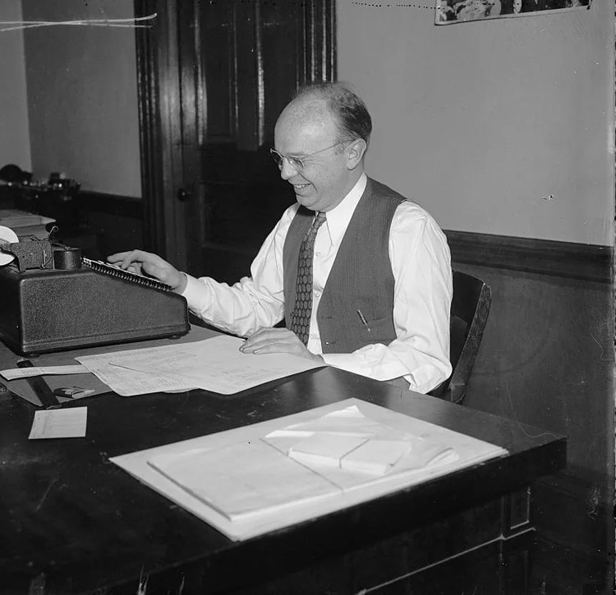 A man in a vest runs an adding machine over a sheet of figures on a dark desk.
