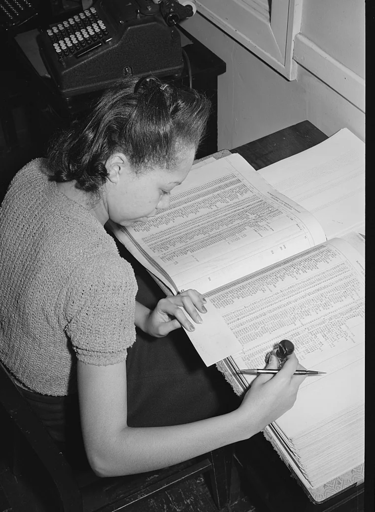 A bookkeeper follows a line in an open ledger, with an adding machine on the desk behind her.