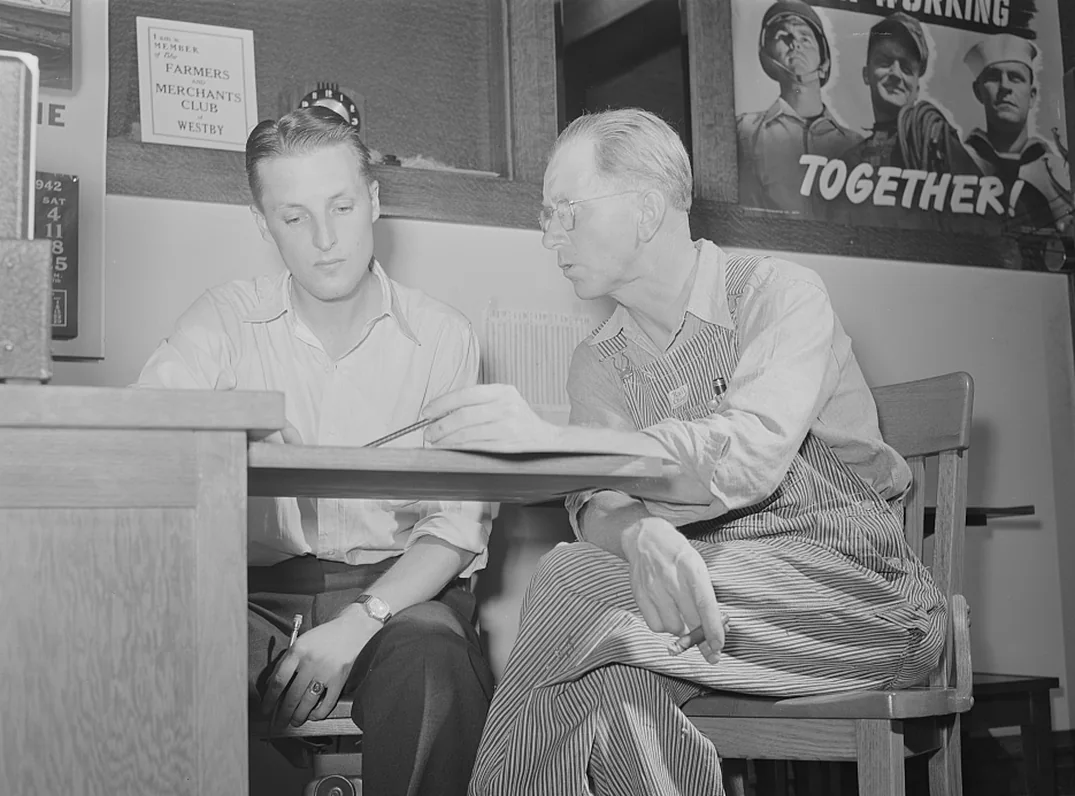 Two men sit at a desk and look at one sheet. A club sign hangs on the wall behind them.