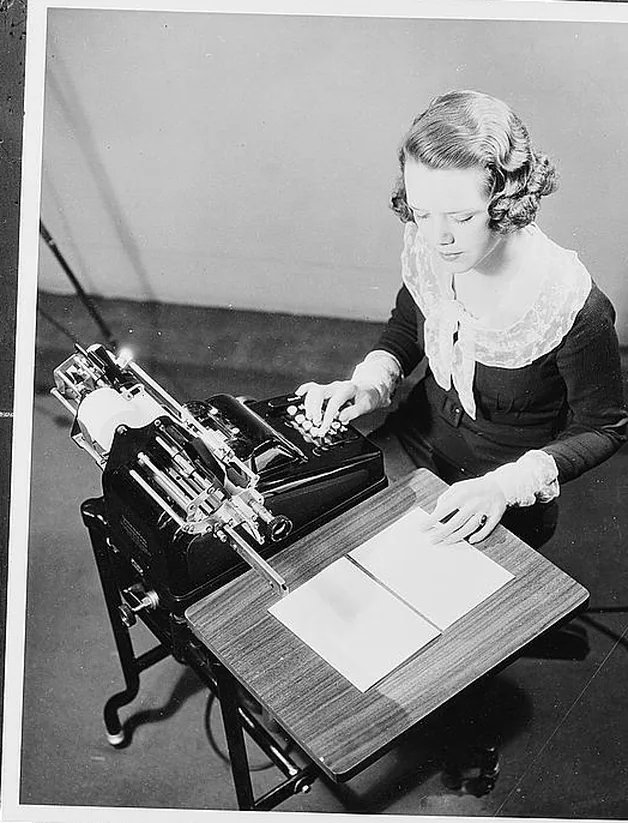 A woman runs an adding machine. The sheet on the stand in front of her is blank.