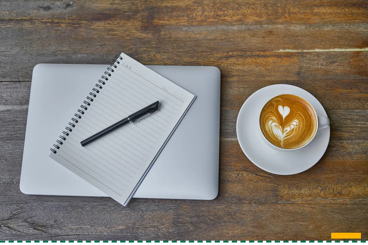 A spiral notepad and a pen on a laptop beside a cup of coffee