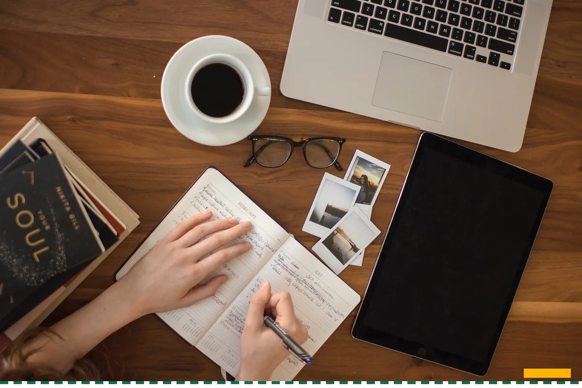 An owner writing in a notebook at a wooden table beside a coffee, a laptop and a tablet