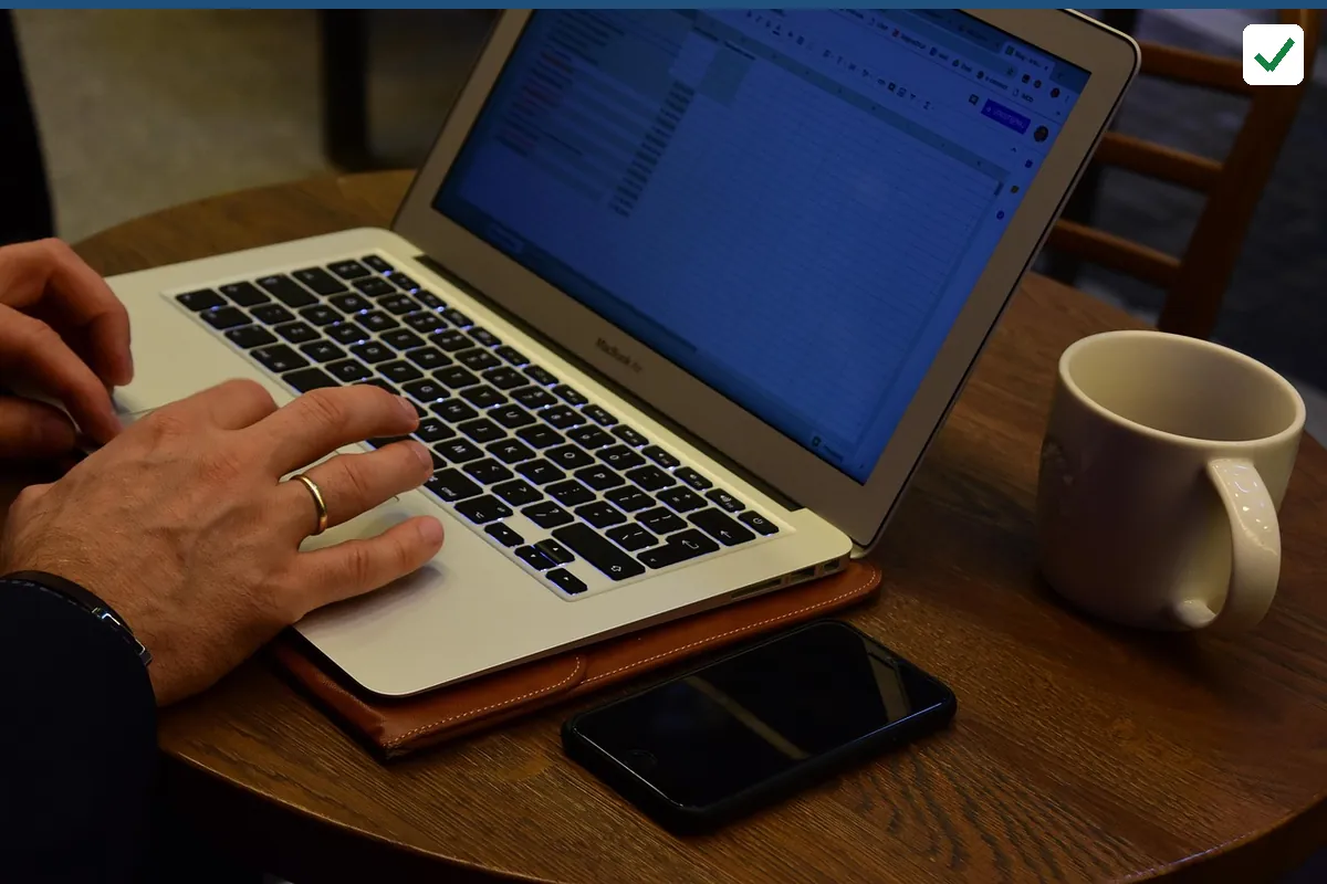 Hands typing on a laptop on a wooden table beside a mug and a phone