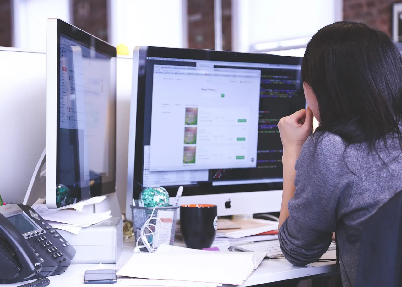 A person at two monitors with papers and a phone