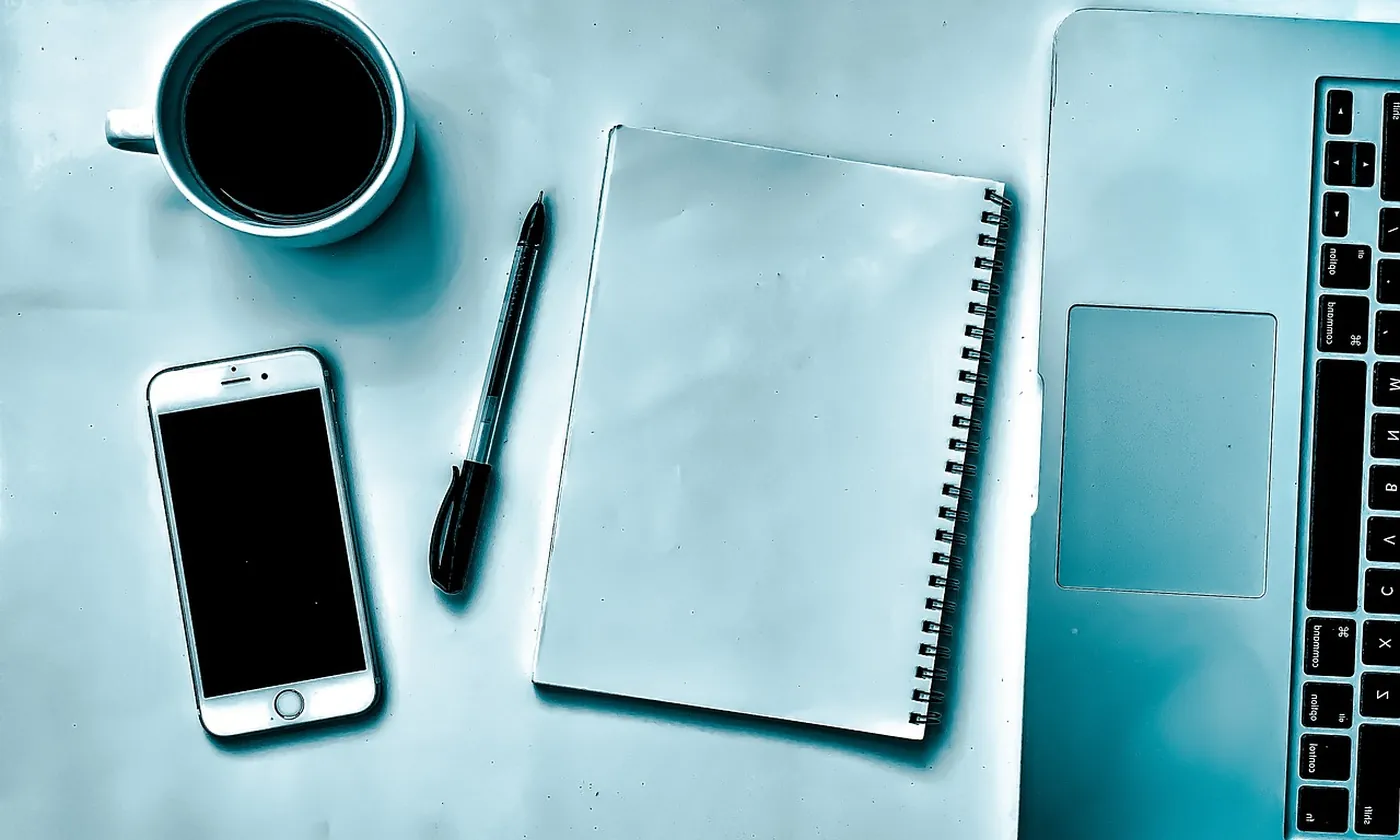 A laptop, a blank notebook, a phone, and coffee on a work table