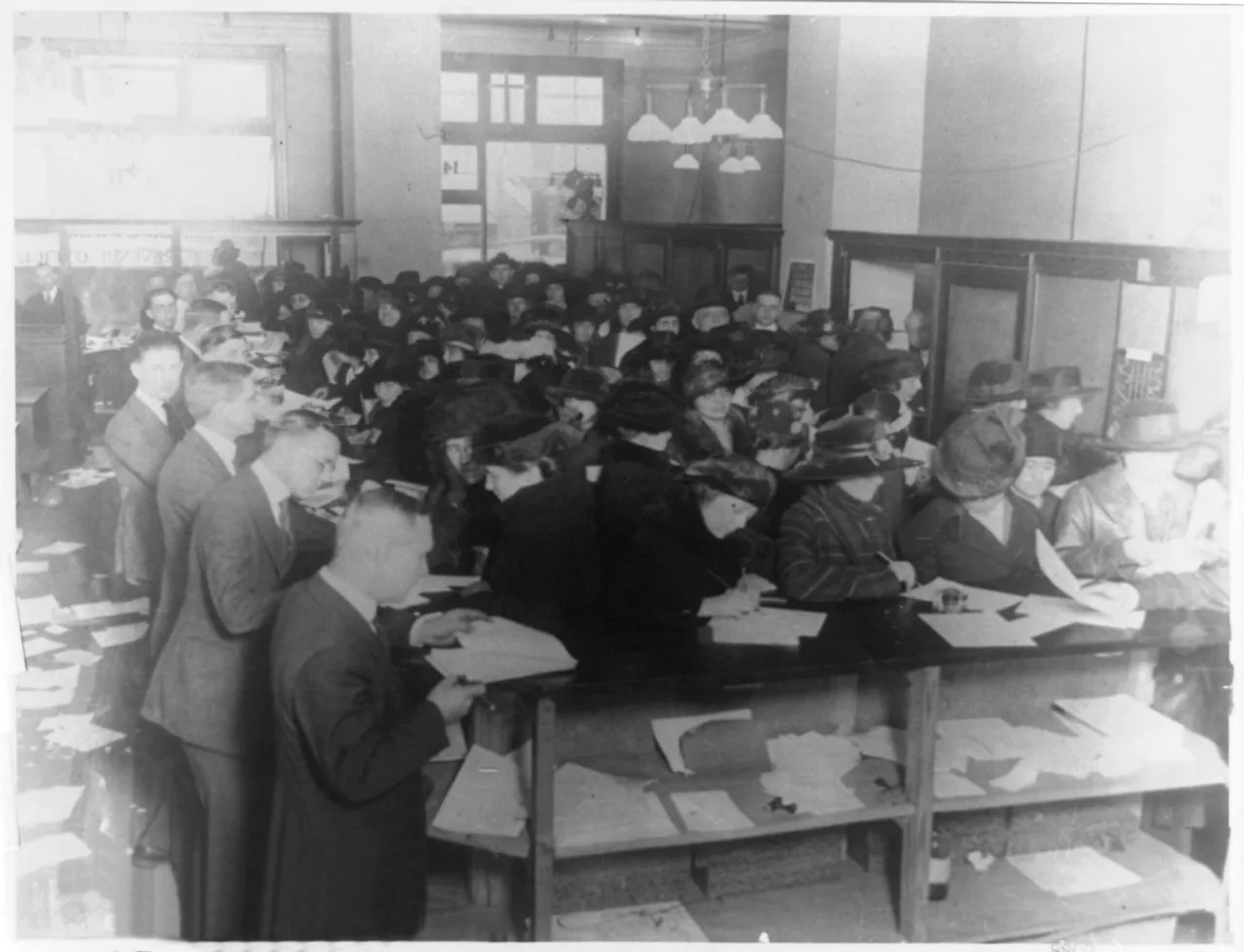 A crowded room of people standing and sitting at long tables, each one writing on a paper form