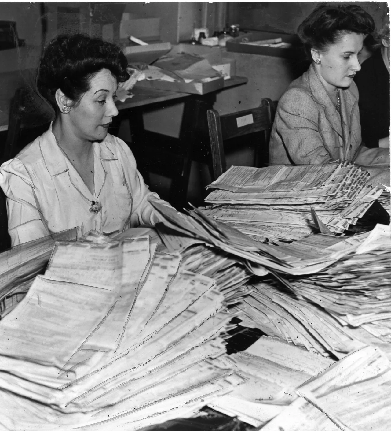 Two women sit at a desk buried in stacks of returns, one holding a sheaf and the other looking at the pile