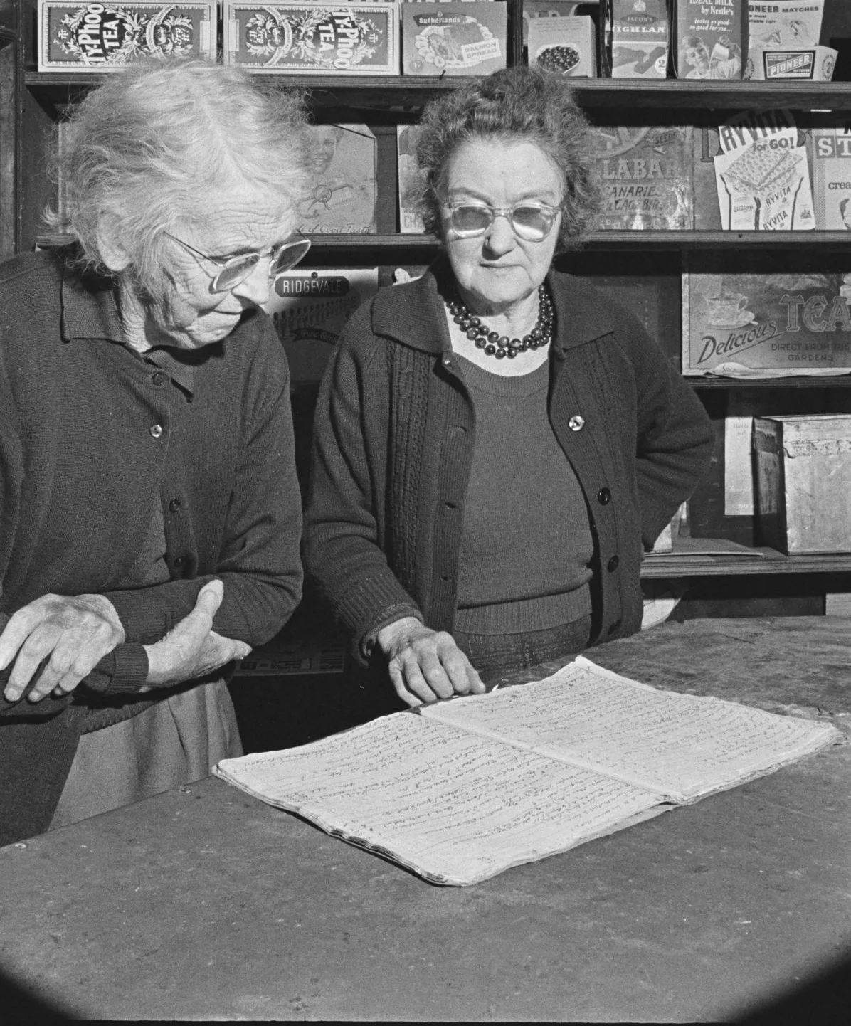 Two shopkeepers stand at the counter after close, one pointing at an open account book