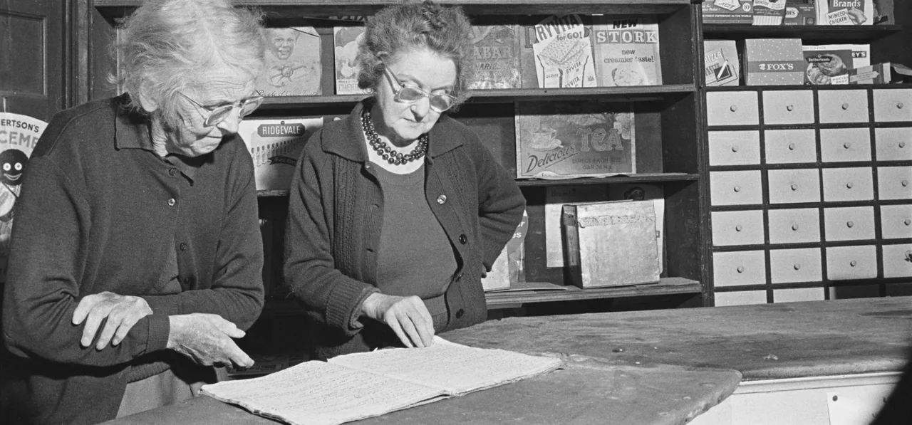 Two shopkeepers look down together at one open account book on the counter between them