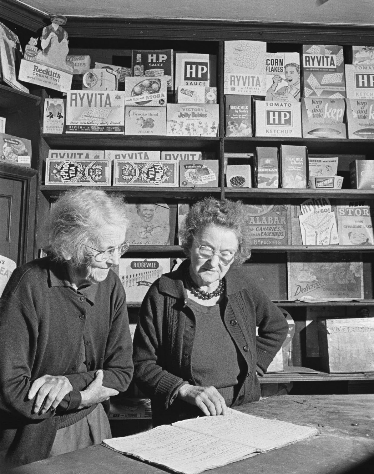 Two shopkeepers read an open account book on the counter, with stocked shelves behind them