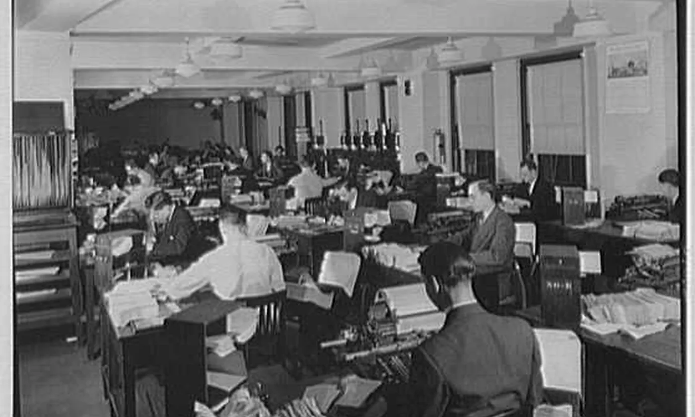 A large bookkeeping floor, with people at desks and stacks of paper between them.