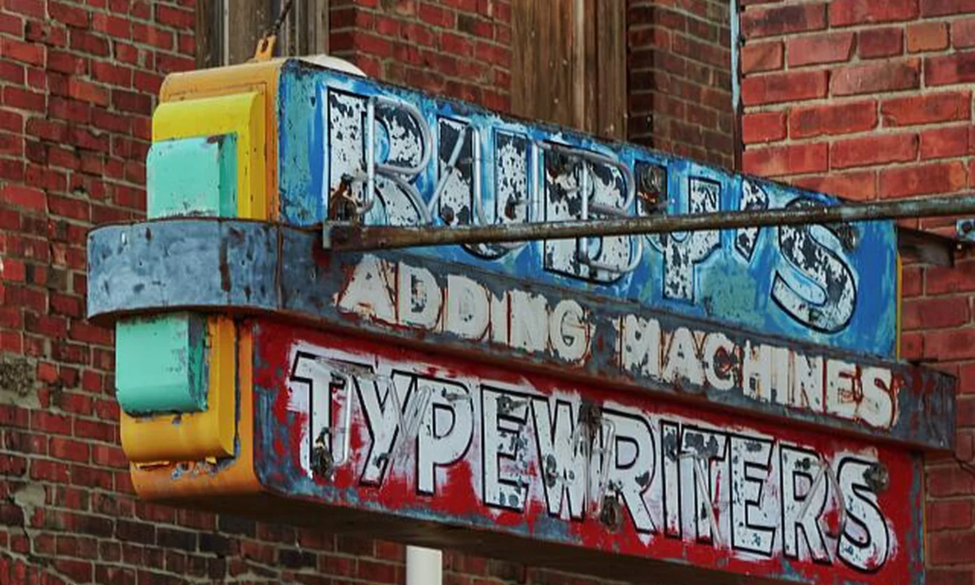 A painted shop sign for adding machines and typewriters, fixed to a brick wall.
