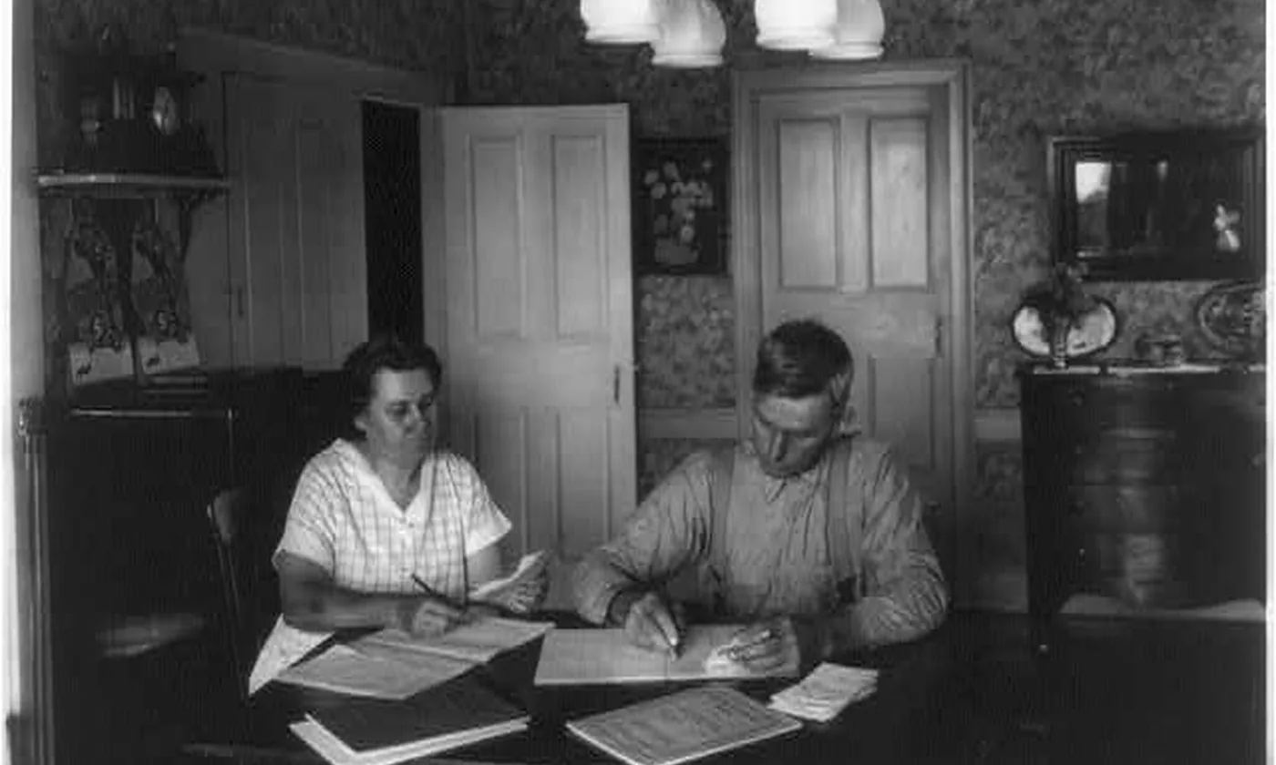 A couple at a dining table, each writing on a sheet, with more papers stacked in front of them.
