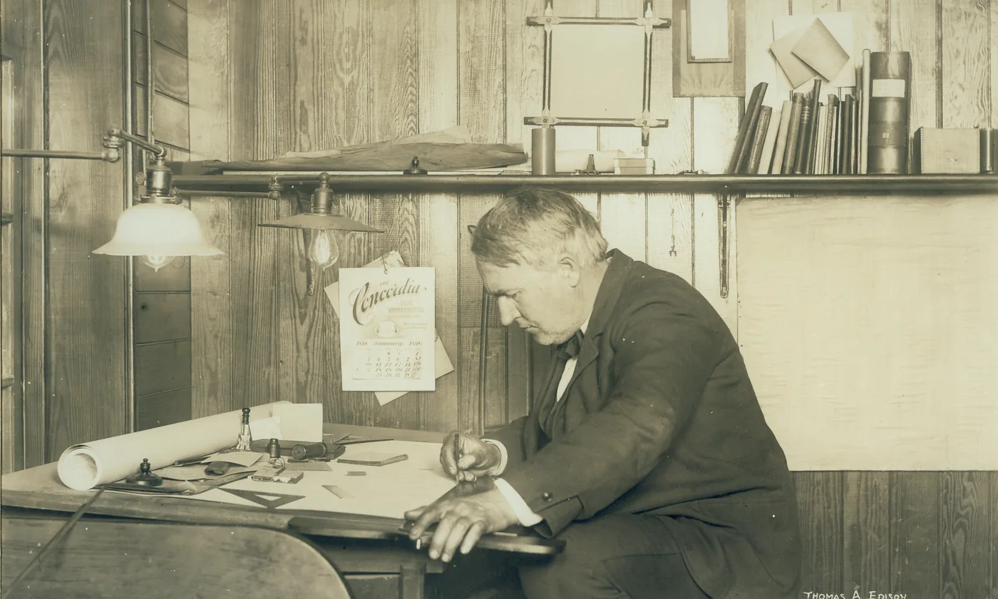 A man writes at a wooden desk in 1905, with a January wall calendar and papers under the writer's hands.