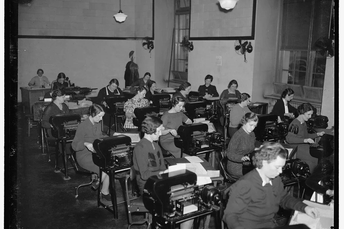 Women at adding machines work through stacks of paper in a tax office.