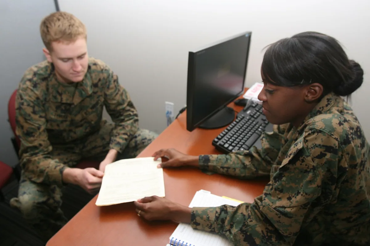 Two people at an office table reviewing a printed form, a monitor and keyboard beside them.