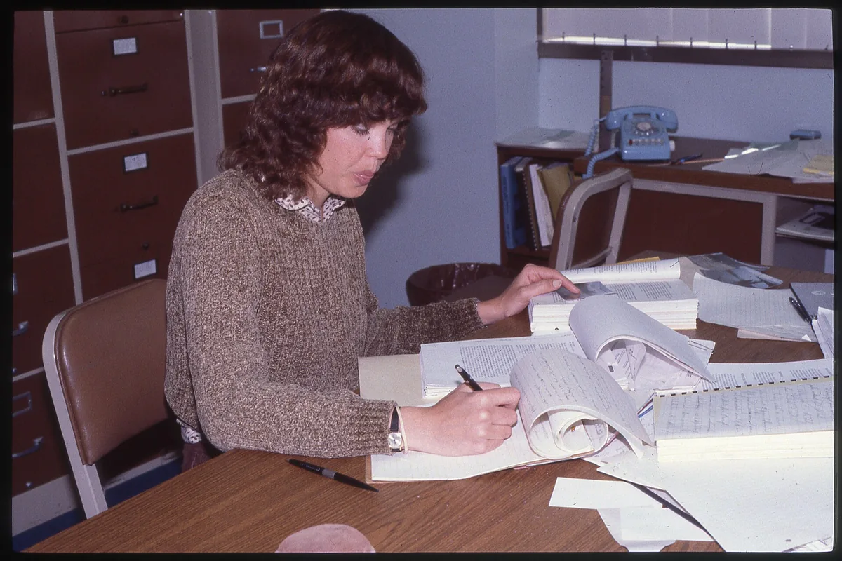 A person writing in a large open ledger at a wooden table, with stacks of papers and file drawers behind.