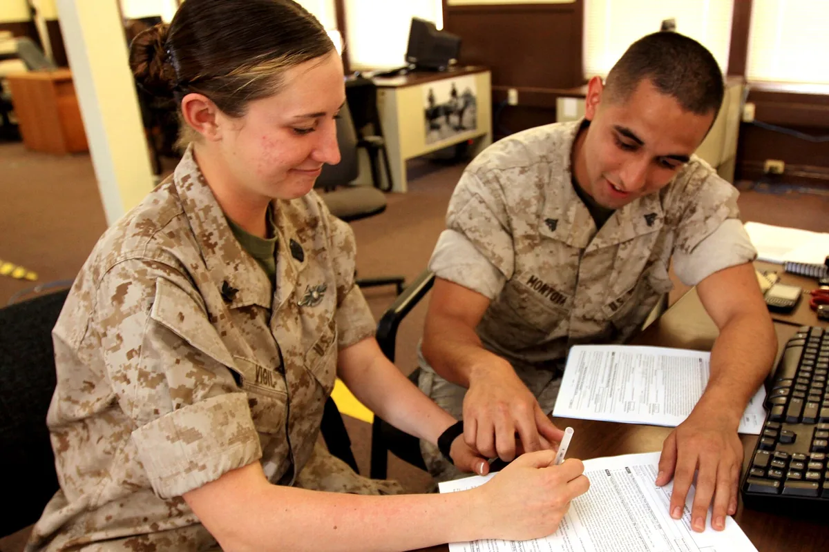 Two people at an office table, one writing on a form while the other points at the page.