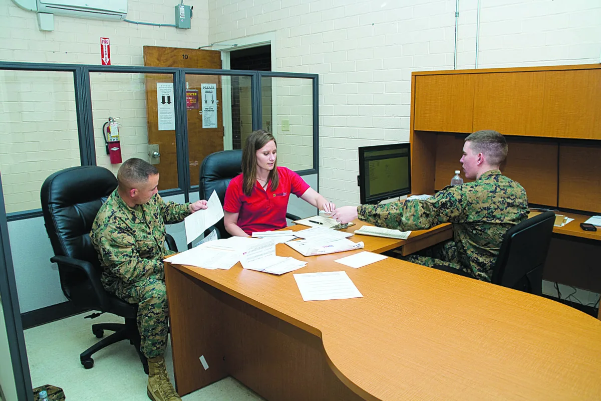 Three people around an office table covered with papers, passing a page across.