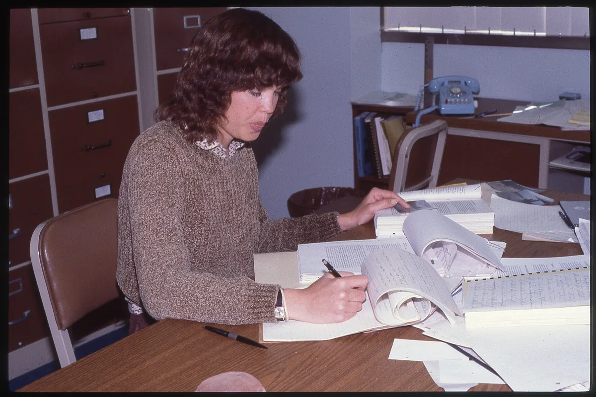 A woman writes in an open ledger while stacks of papers sit on the table.