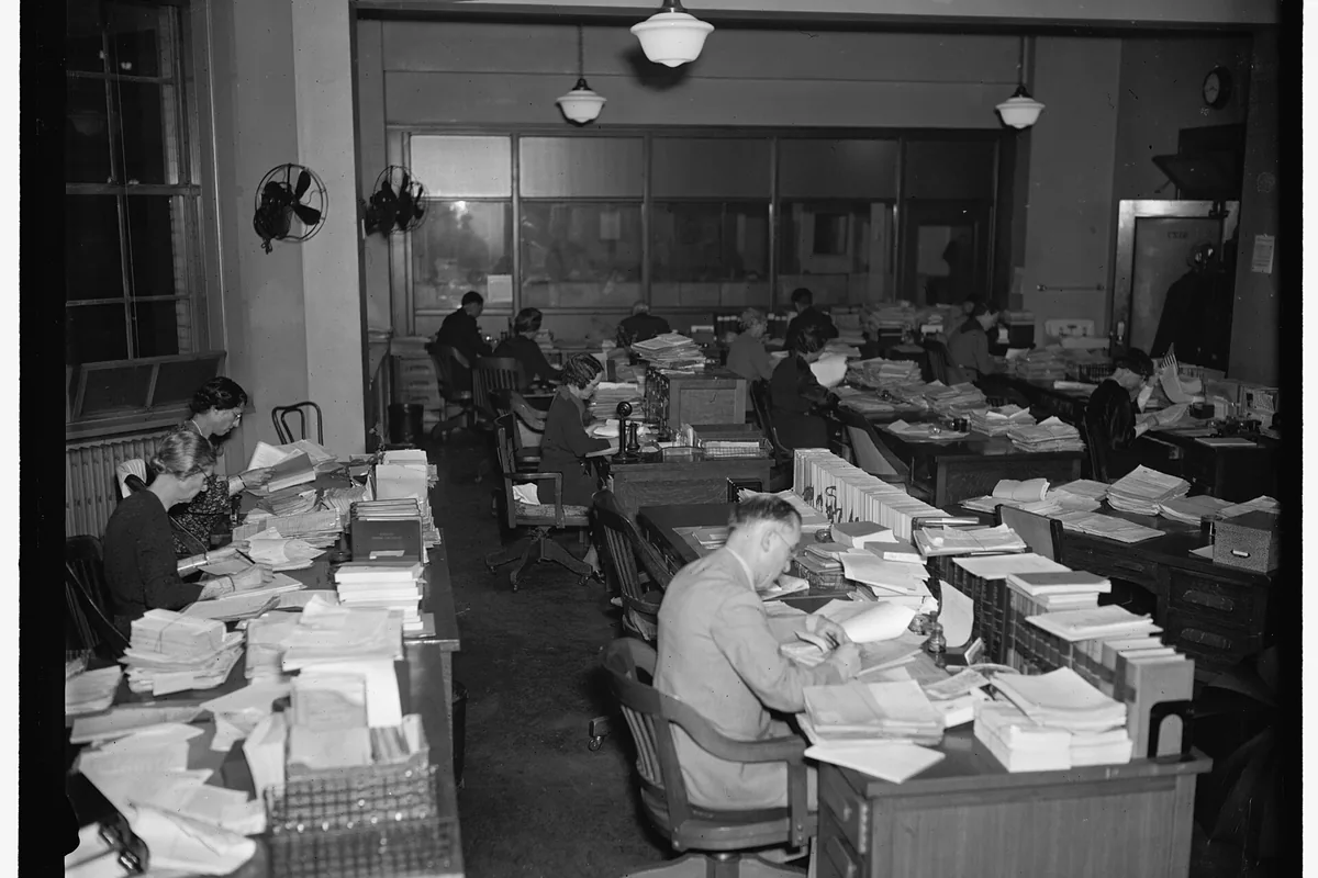A man reads a thick stack of papers at a crowded office table.