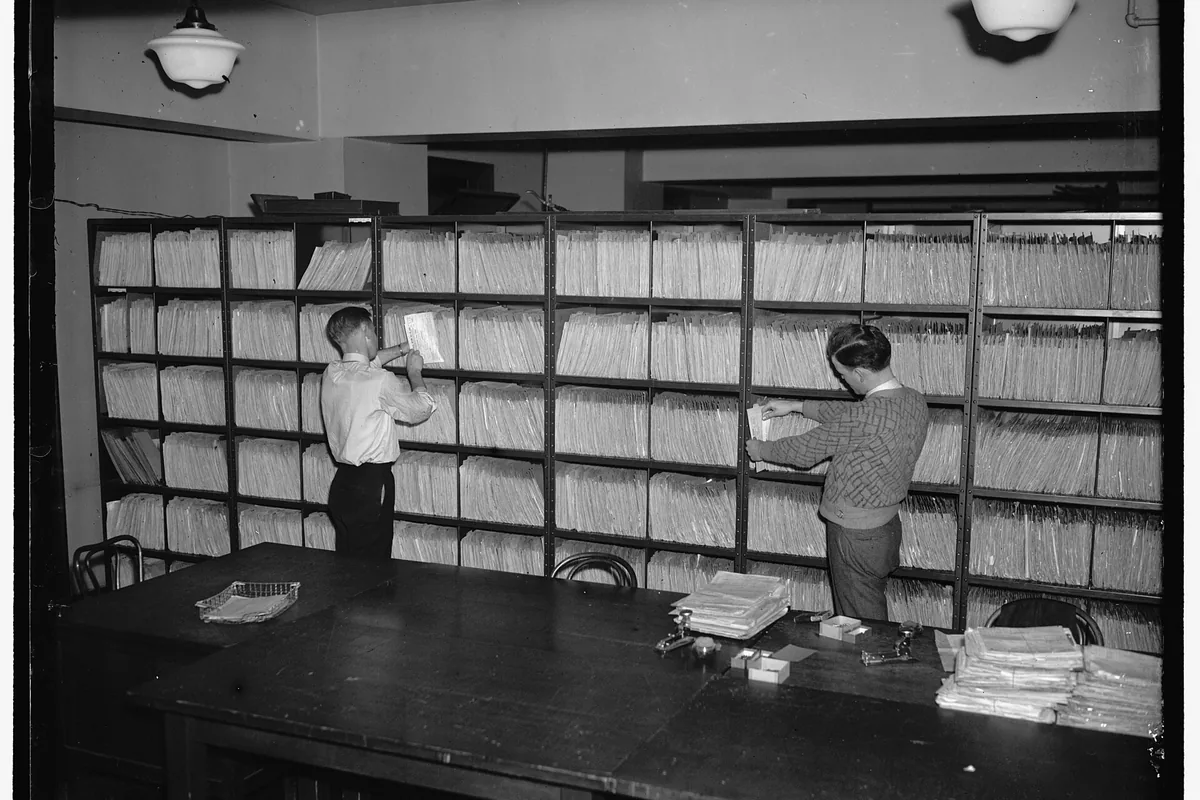 Two men pull folders from shelves packed with files.