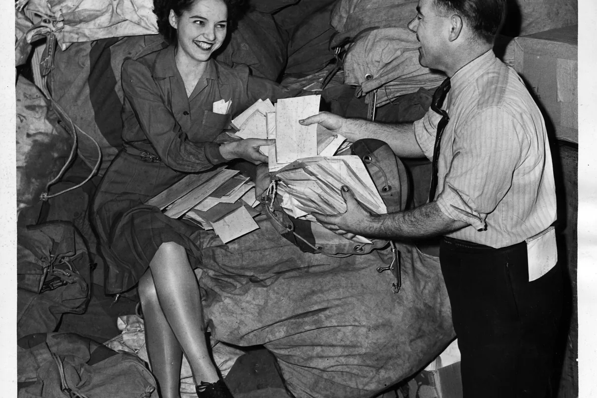 A woman and a man sort a tall stack of tax forms beside mail sacks.