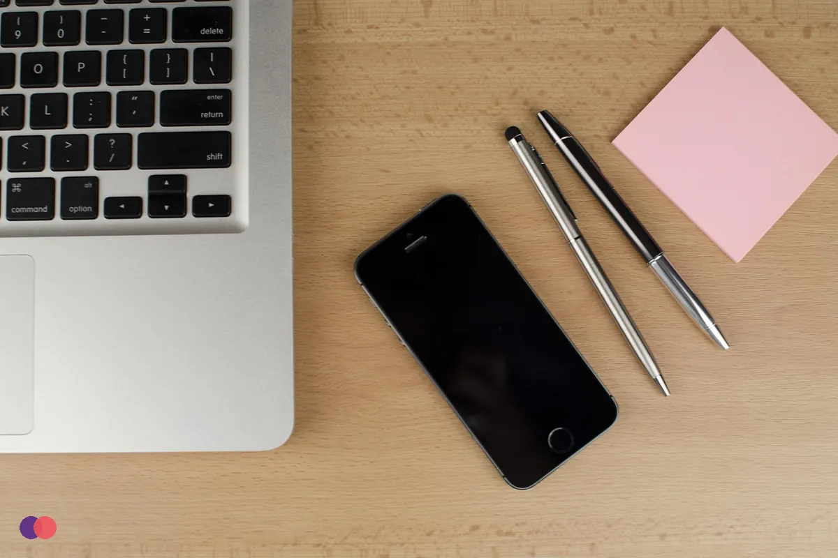 A blank pink sticky note beside a laptop, a phone and two pens