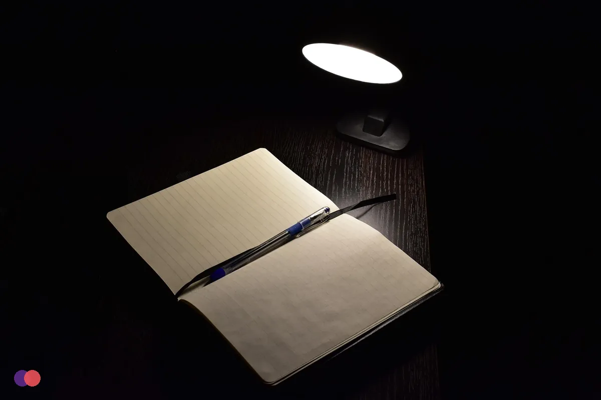 A lamp lighting an open notebook and a pen late in the evening