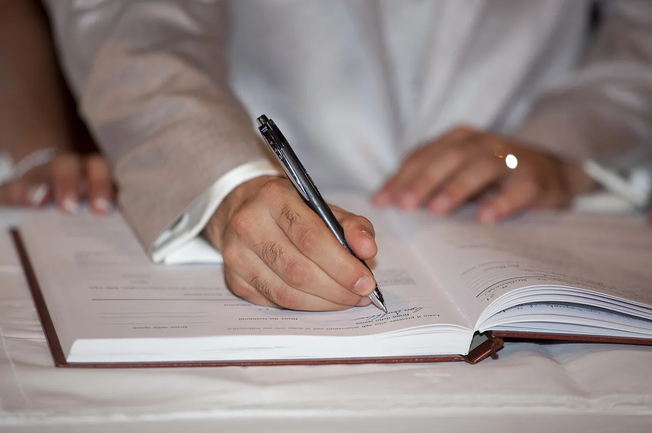 A hand signs an open register with a pen on the page