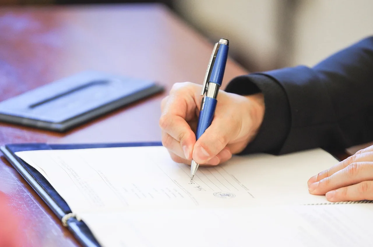 A hand holds a blue pen and signs a page in a dark folder