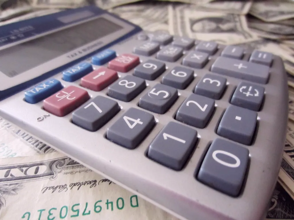 A calculator with tax keys rests on United States currency, including a one hundred dollar bill.