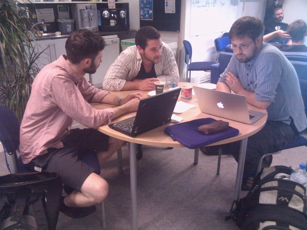 Three people sit at a round table with two laptops and a sheet of paper, talking through a problem.